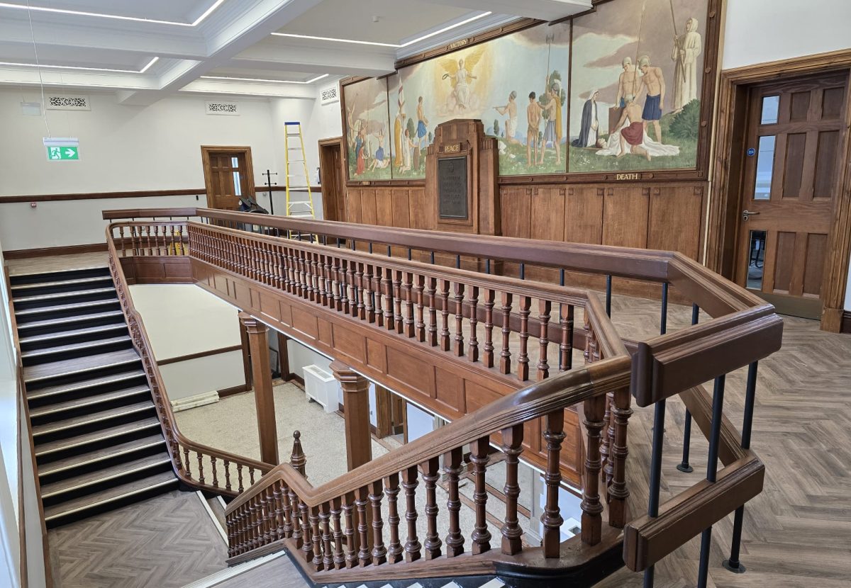 Elevated view looking down a wooden staircase banister towards a upper-level balcony corridor, featuring classical painted mural artwork mounted along the wall.