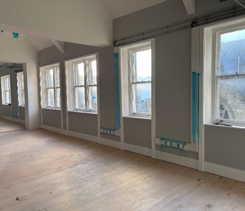 A large, multi-paneled stained glass bay window set within a white arched alcove inside a building under construction, featuring decorative heraldic designs and a dark ornate wooden ceiling tracery above.